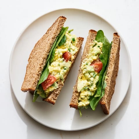 Golden avocado egg salad sandwich layered with fresh spinach and ripe tomato slices on toasted whole grain bread.  