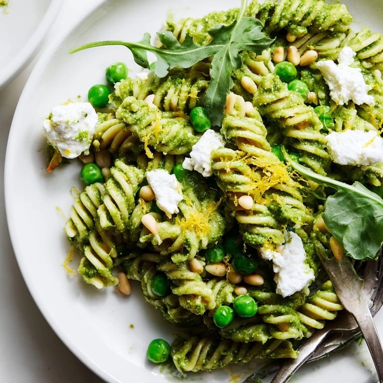 Spoon a serving of Spring Green Pesto Pasta Salad into a white bowl, revealing creamy pesto coating and bright green peas and arugula.